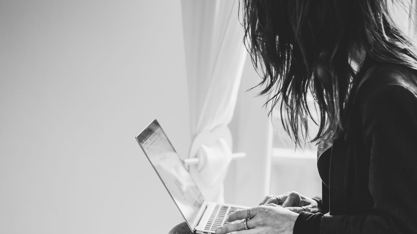 Tiziana di profilo, con i capelli sul viso, scrive al portatile seduta accanto alla finestra.
